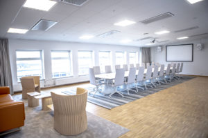 En bild på ett modernt konferensrum med bekväma stolar runt ett stort bord. På bordet ligger block och pennor redo för anteckningar. Rummet är ljust med stora fönster och modern belysning. En LED-skärm hänger på väggen och en projektor är monterad i taket. På ena sidan av rummet finns en whiteboard för att skissa upp idéer. Konferensrummet ligger i Borås Kongress, en plats som präglas av modernitet, funktionalitet och tillgänglighet. Perfekt lokal för att boka ditt möte.